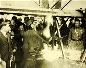 A woman in a dress breaks a bottle of champagne against the fuselage of an airplane. A man in a pilot's uniform on the other side of the plane squints as the champagne sprays him in the face